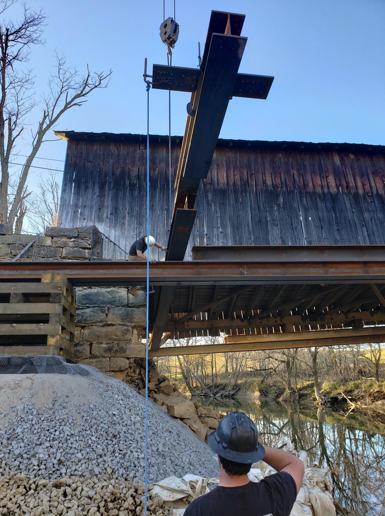 Worker guiding steel cross beam lifted by crane over Grange City covered bridge abutment